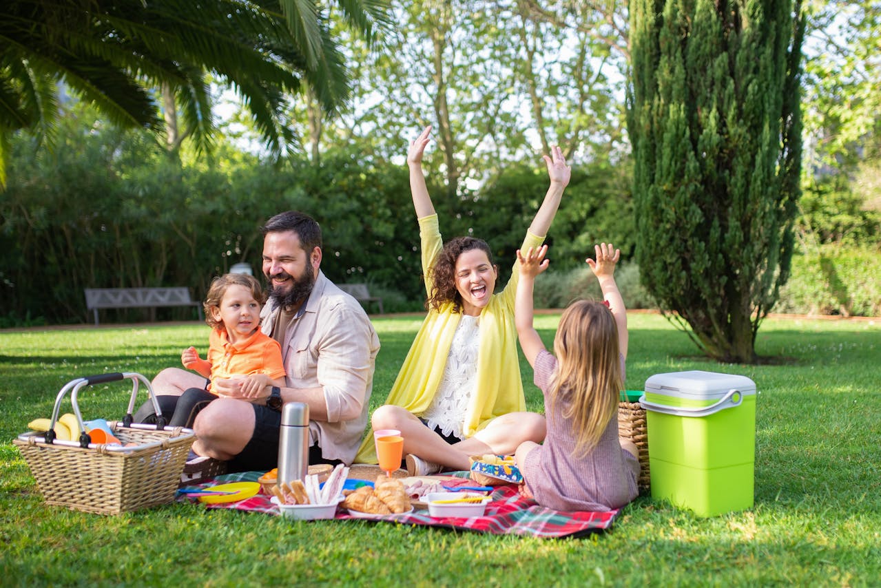 Happy family having a picnic on a blanket in a sunny park, surrounded by food, drinks, and trees