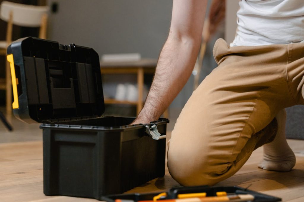 Man kneeling on wooden floor, reaching into black plastic toolbox, tool tray with hand tools nearby, casual indoor setting