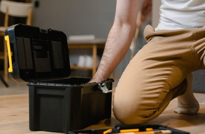 Man kneeling on wooden floor, reaching into black plastic toolbox, tool tray with hand tools nearby, casual indoor setting