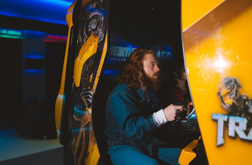 Man with long red hair playing an arcade racing game, seated in a yellow game cabinet, focused expression, neon lights in background