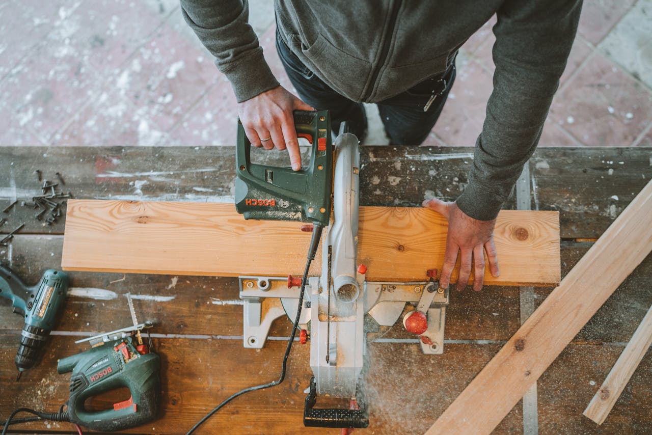 Person using an electric saw, cutting a wooden board on a miter saw, workshop setting, tools scattered on workbench, construction or carpentry in progress
