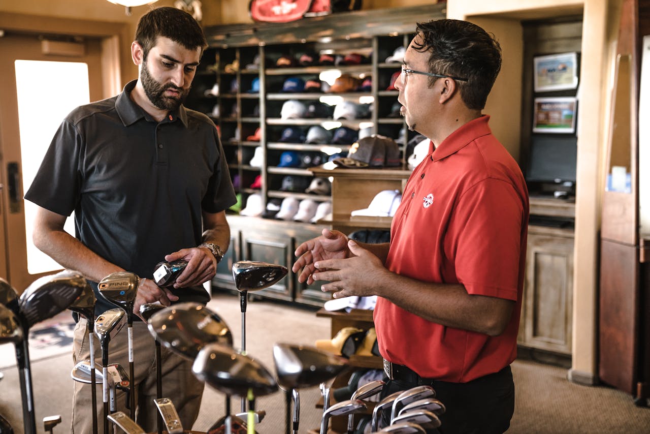 Two men discussing golf clubs in a pro shop, surrounded by clubs and shelves of hats