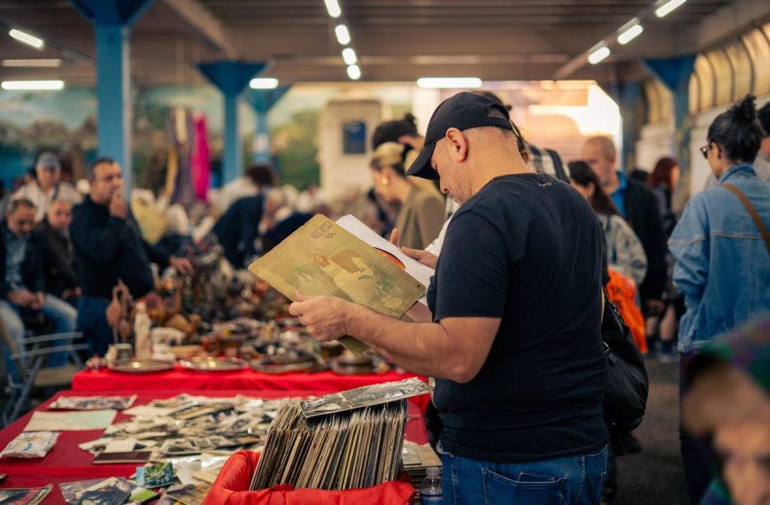 Man in black cap browsing vintage vinyl records at a busy indoor flea market, surrounded by antique items and shoppers