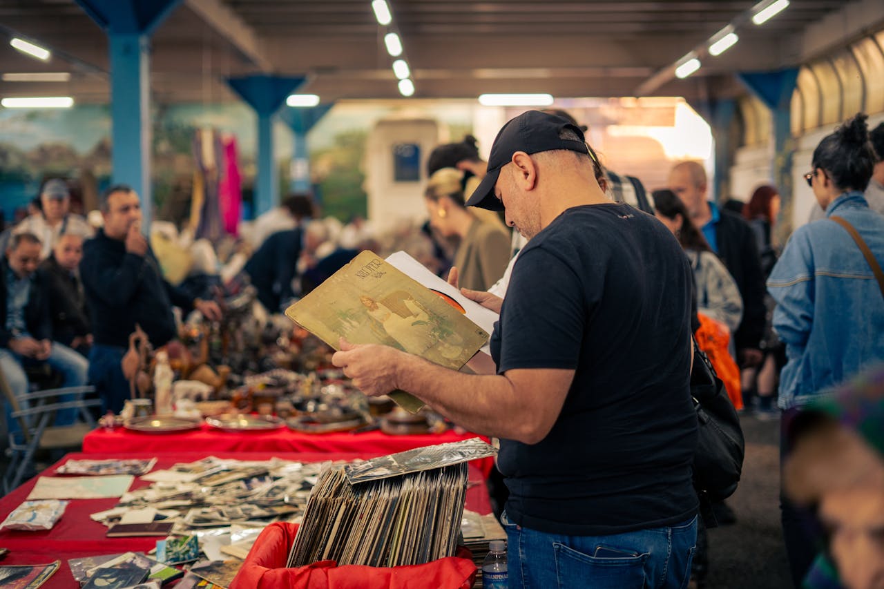 Man in black cap browsing vintage vinyl records at a busy indoor flea market, surrounded by antique items and shoppers