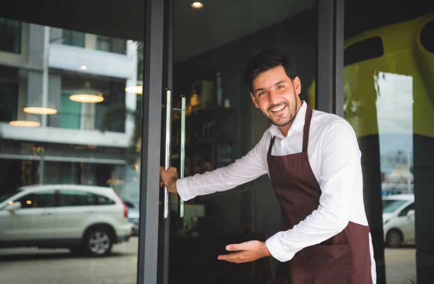 Smiling man in apron opening a glass door, welcoming customers into a shop or café