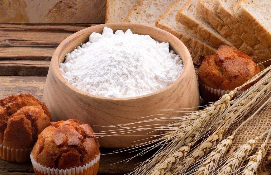 Bowl of white flour, surrounded by wheat stalks, round bread rolls, and sliced bread, displayed on a rustic wooden surface, representing baking ingredients and products