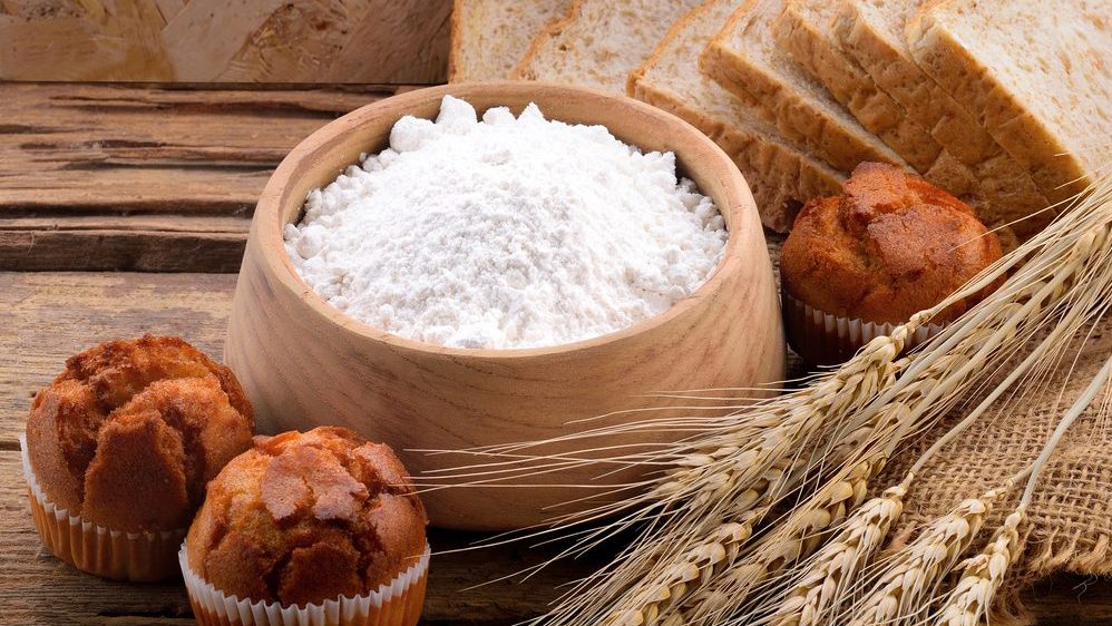 Bowl of white flour, surrounded by wheat stalks, round bread rolls, and sliced bread, displayed on a rustic wooden surface, representing baking ingredients and products