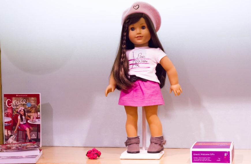 Brunette American Girl doll in a pink skirt, white graphic tee, matching pink beret, and grey ankle boots, standing on a display stand