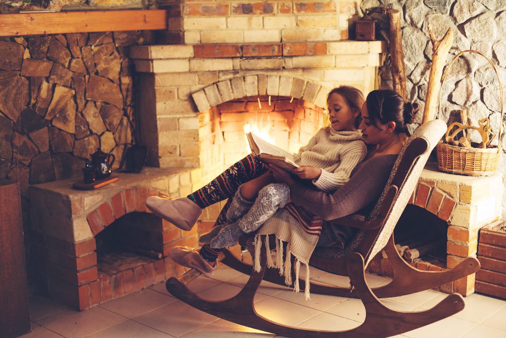 Mother and daughter reading in rocking chair near warm brick fireplace, cozy indoor setting
