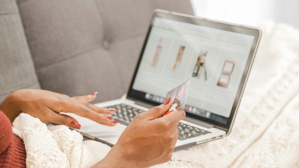 Person holding a credit card, browsing an online shopping website on a laptop, sitting on a sofa, product images visible on screen, relaxed indoor setting