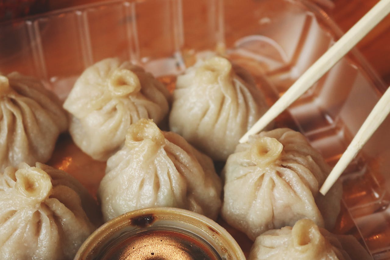 Close-up of soup dumplings in plastic tray, soy sauce container, chopsticks picking one dumpling, soft and steamed texture