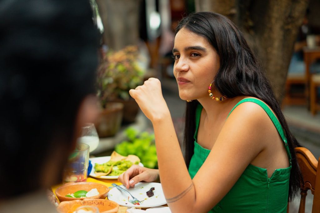 Young woman sitting at an outdoor table, eating and looking to the side, plates with food and drinks on the table