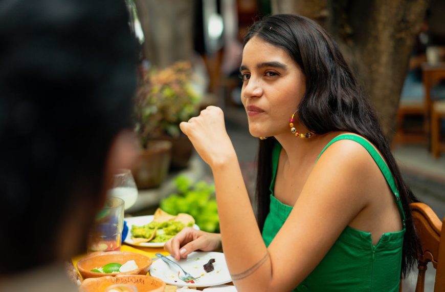 Young woman sitting at an outdoor table, eating and looking to the side, plates with food and drinks on the table