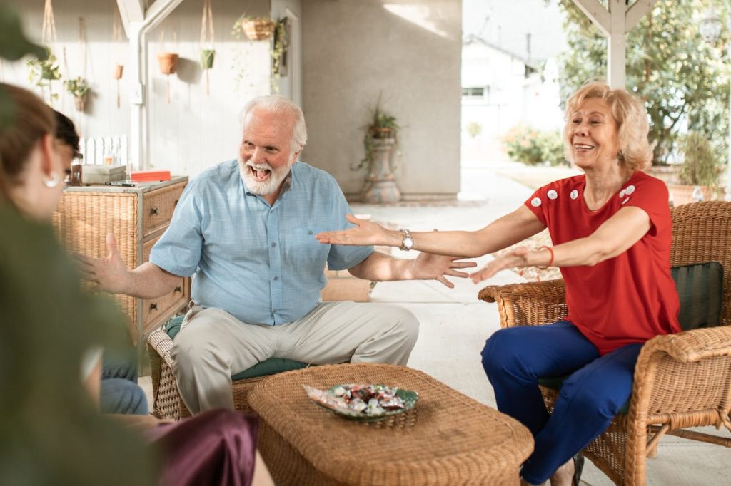 Smiling elderly couple, arms outstretched, sitting on wicker chairs, talking with family on a sunlit porch, cheerful expressions