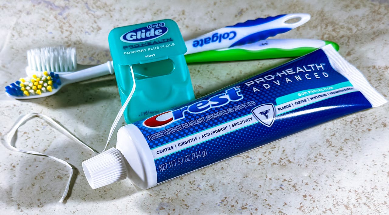 Crest toothpaste tube, Oral-B Glide floss, Colgate toothbrush, loose dental floss, placed on a bathroom countertop