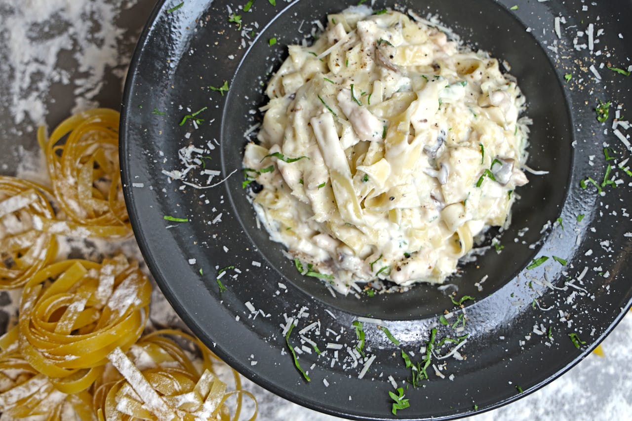 Plate of creamy fettuccine pasta with herbs, mushrooms, and parmesan cheese, surrounded by raw pasta nests dusted with flour