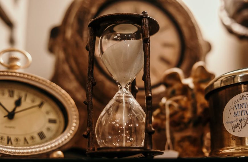 Hourglass with sand, surrounded by vintage clocks and antique collectibles, set on a dimly lit shelf