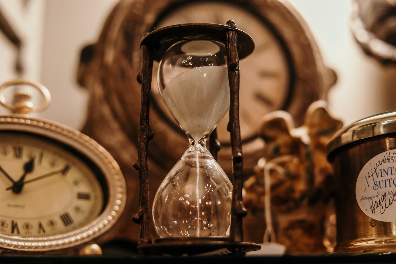 Hourglass with sand, surrounded by vintage clocks and antique collectibles, set on a dimly lit shelf