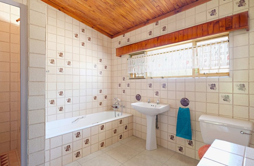 Vintage bathroom with tiled walls, built-in bathtub, pedestal sink, wooden ceiling, large window, blue towel, natural light