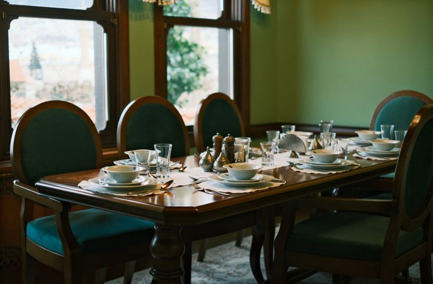 Elegant wooden dining table, six chairs with green upholstery, neatly arranged tableware, bowls, glasses, cutlery, vintage interior with window view