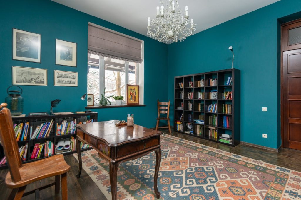 Teal-walled study with chandelier, wooden desk, patterned rug, bookshelf with books, window with roman shade, framed art
