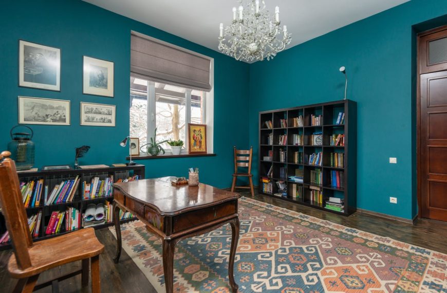 Teal-walled study with chandelier, wooden desk, patterned rug, bookshelf with books, window with roman shade, framed art