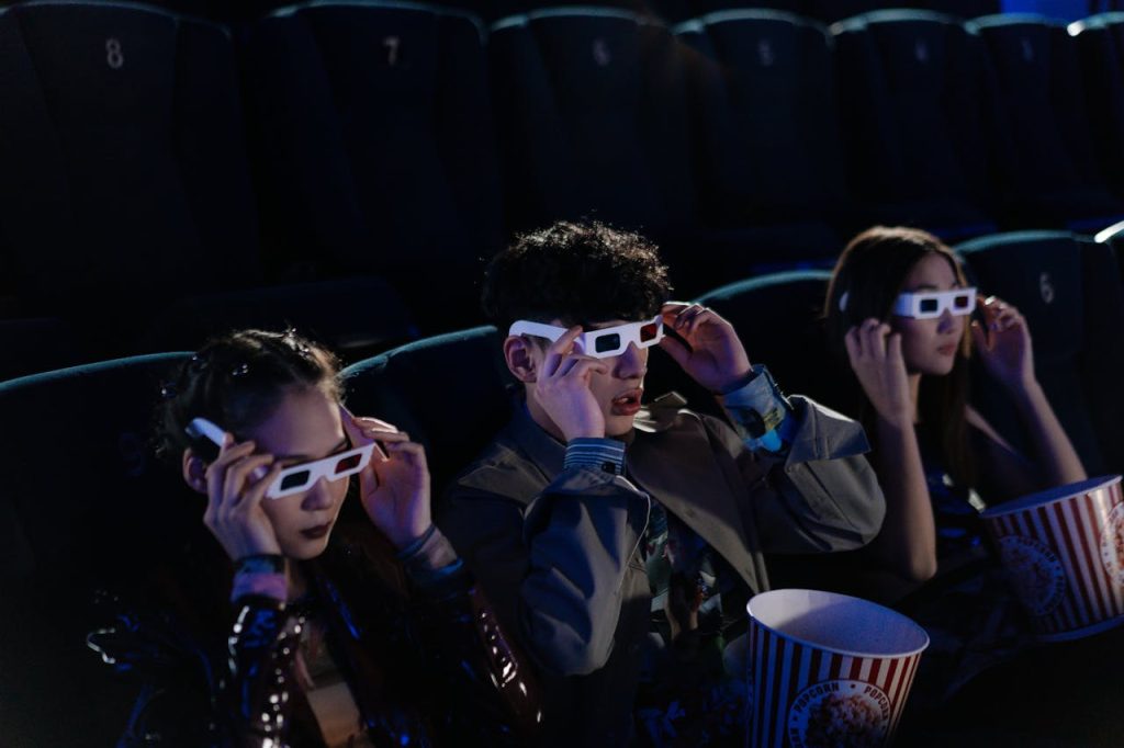 Three people wearing 3D glasses, seated in a dark movie theater, holding popcorn buckets, watching a film attentively
