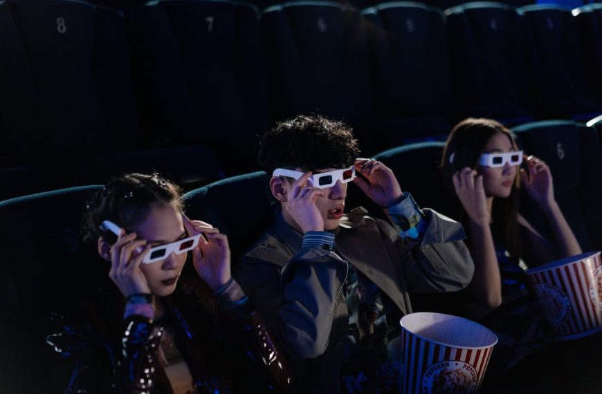 Three people wearing 3D glasses, seated in a dark movie theater, holding popcorn buckets, watching a film attentively