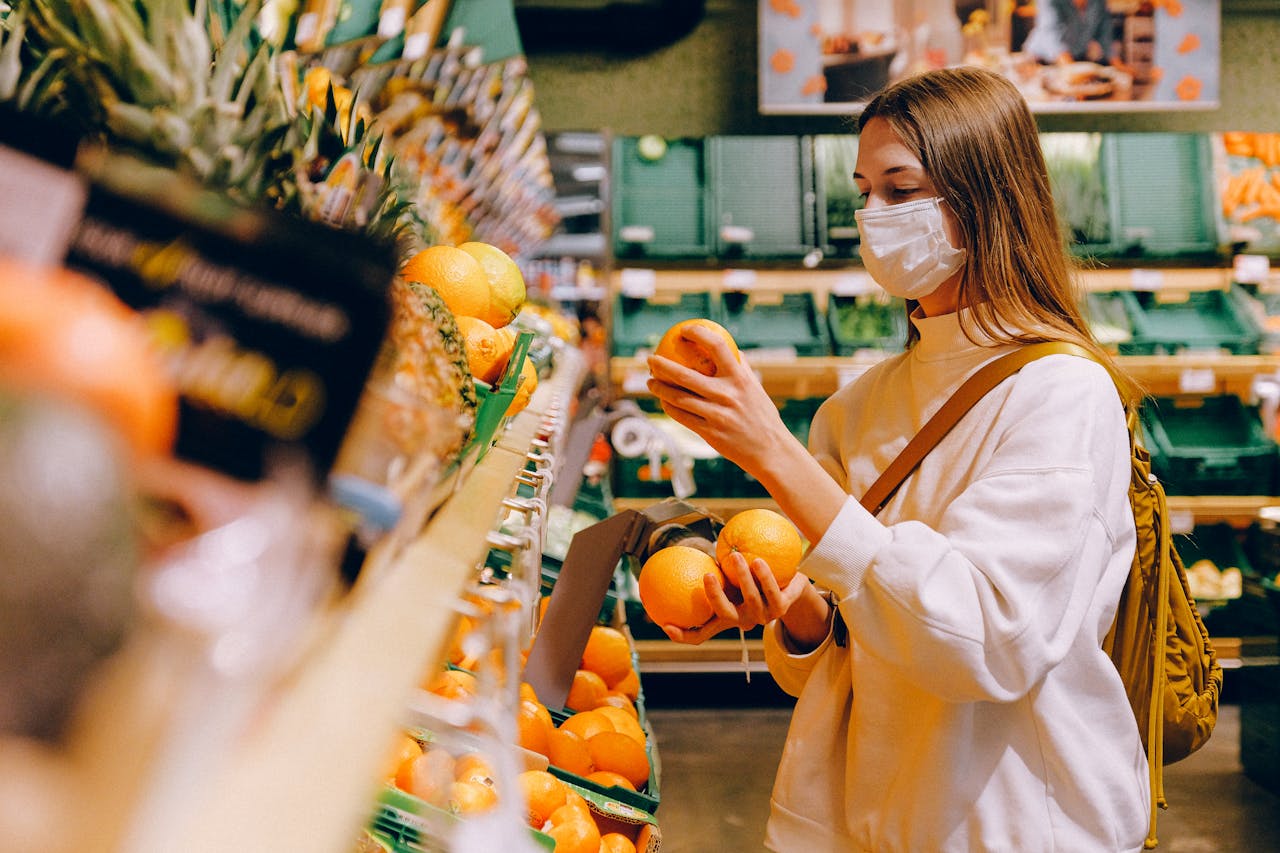 Woman wearing face mask, holding oranges, shopping in a grocery store produce aisle, wearing white sweater and backpack