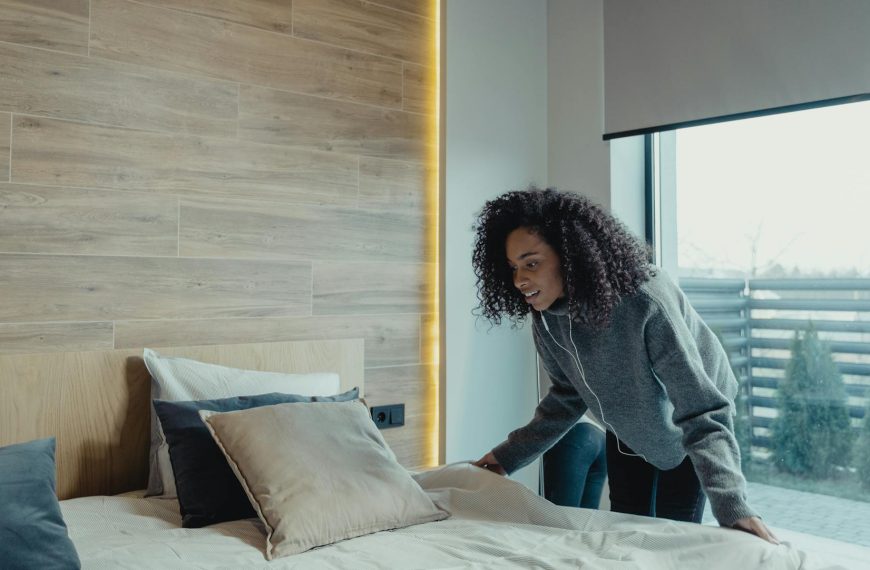 Woman making a bed, modern bedroom setting, neatly arranged pillows, large window with city view, natural light filling the room, minimal and clean decor