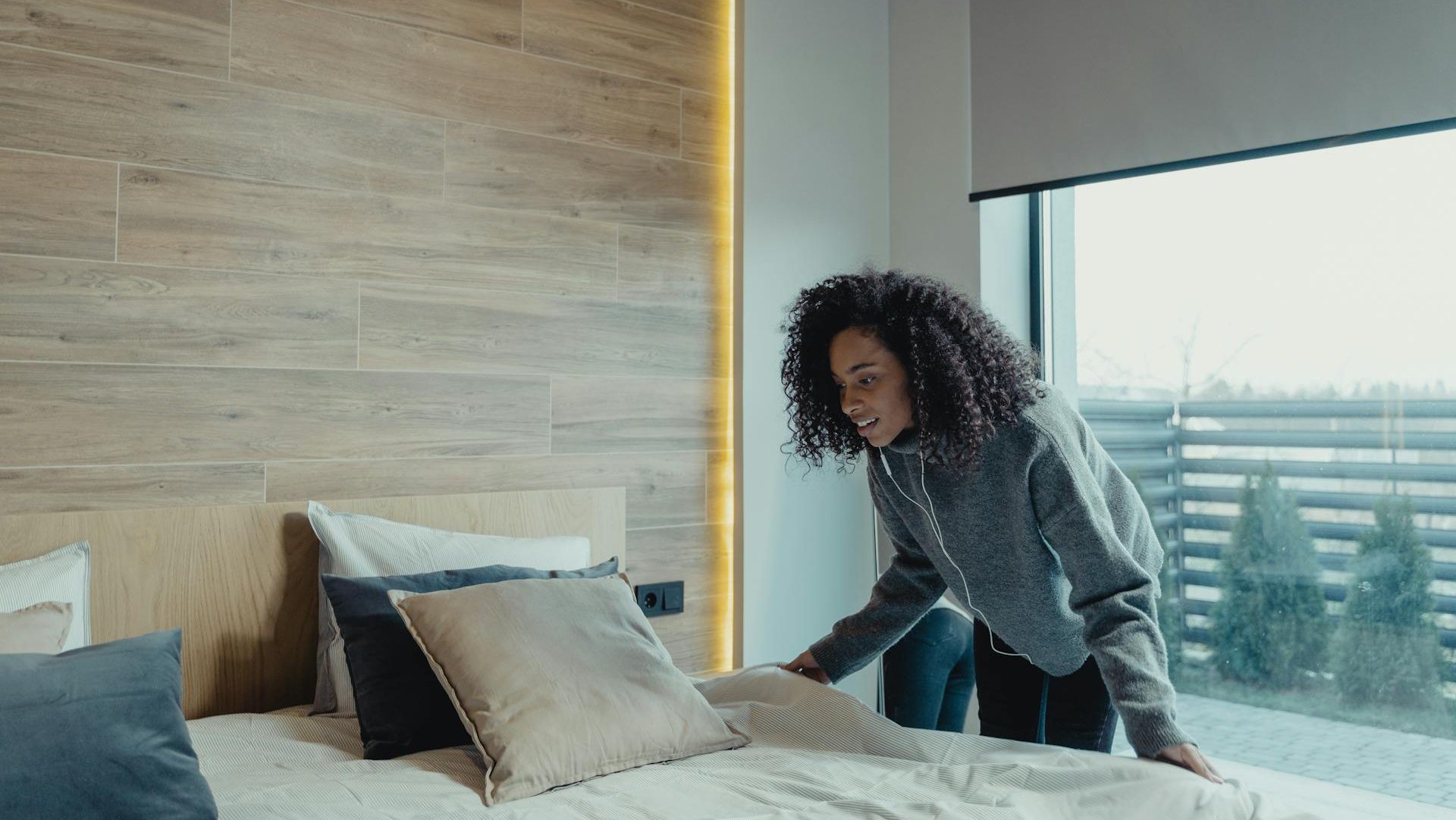 Woman making a bed, modern bedroom setting, neatly arranged pillows, large window with city view, natural light filling the room, minimal and clean decor