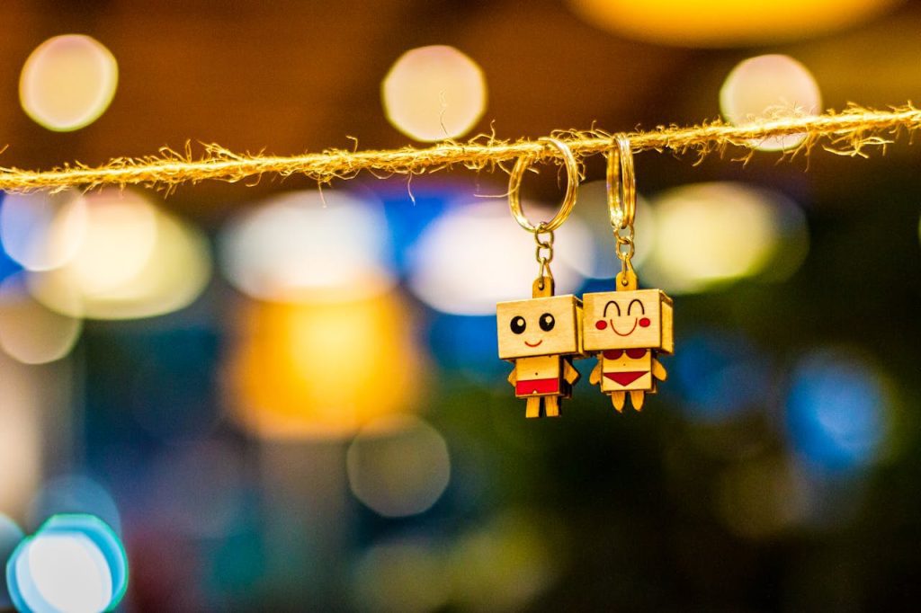 Two wooden keychains with cartoon faces, hanging on a rope, colorful blurred lights in the background, handcrafted accessories