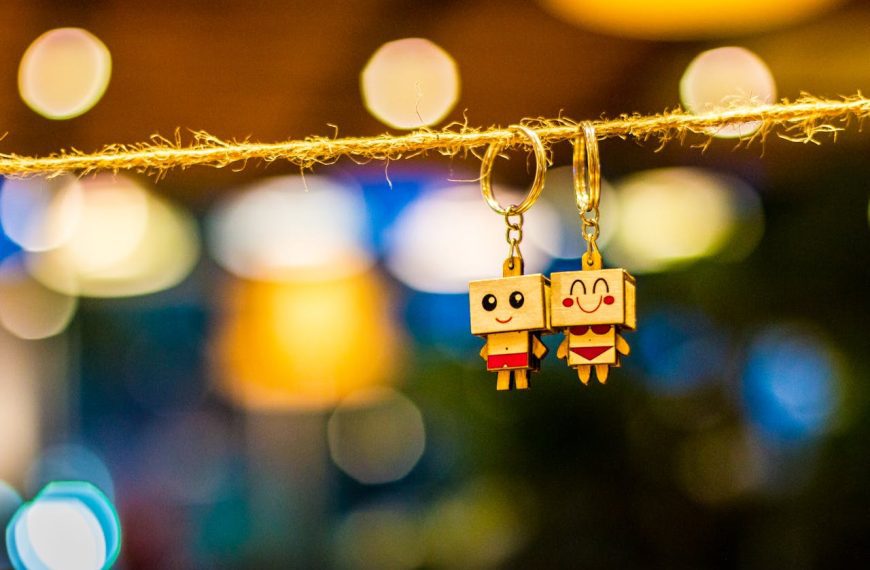 Two wooden keychains with cartoon faces, hanging on a rope, colorful blurred lights in the background, handcrafted accessories
