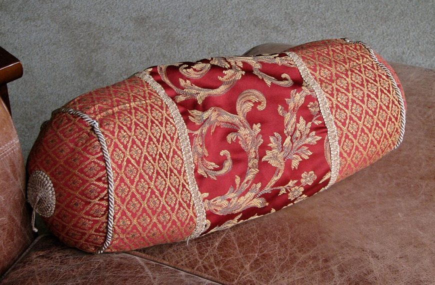Decorative cylindrical pillow, traditional woven pattern, red and gold fabric, placed on a textured surface, used for back or neck support