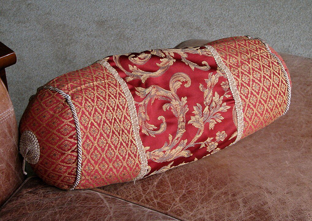 Decorative cylindrical pillow, traditional woven pattern, red and gold fabric, placed on a textured surface, used for back or neck support