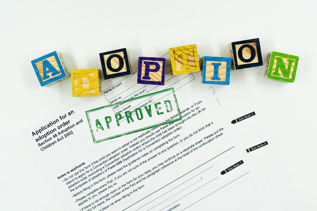 Colorful blocks spelling “ADOPTION” on a white background, stamped document underneath with the word “APPROVED” in green, official paperwork with blank signature lines, symbolizing legal adoption approval