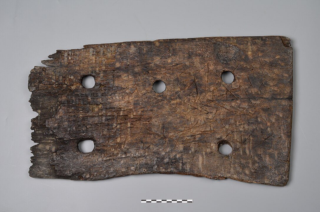 Old, weathered wooden plank, six evenly spaced round holes, rough texture, dark brown coloration, signs of age and decay, possibly part of a historical structure or artifact, displayed on a plain surface