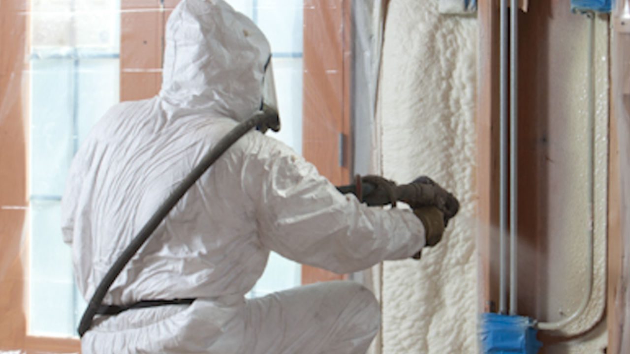 Worker in a white protective suit spraying foam insulation onto open wall studs, interior construction scene.