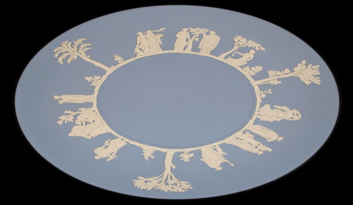 Round Wedgwood ceramic plate, pale blue base, decorated with white neoclassical reliefs of human figures and trees arranged in a circular pattern around the edge