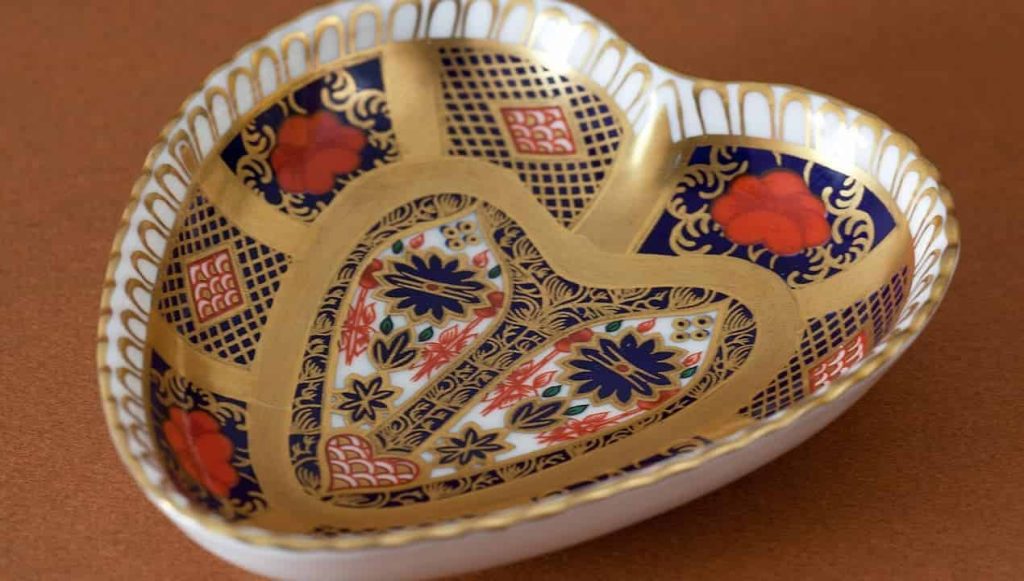Heart-shaped porcelain dish, intricate gold detailing, deep blue and red floral and geometric patterns, glossy finish, resting on a warm brown surface