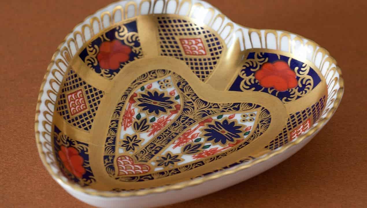Heart-shaped porcelain dish, intricate gold detailing, deep blue and red floral and geometric patterns, glossy finish, resting on a warm brown surface
