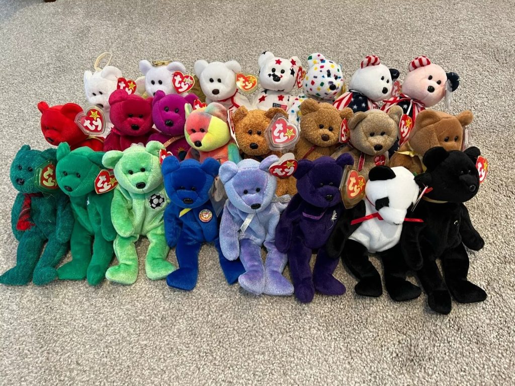 Colorful collection of teddy bears, arranged in rows on a concrete surface, includes various colors like red, green, blue, purple, brown, black, and pastel shades, some bears have accessories like bows or shirts, plush toys displayed for sale or collection