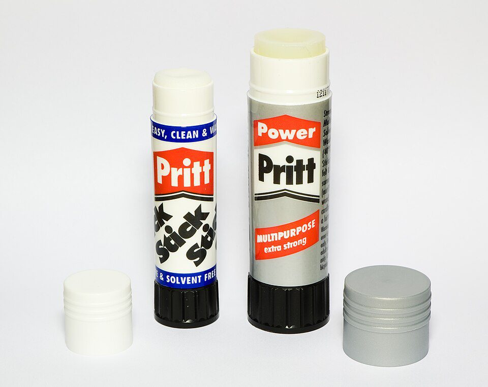 Two glue sticks standing upright, one with a blue label and the other with a red label, caps placed beside them, white background, clear product branding visible