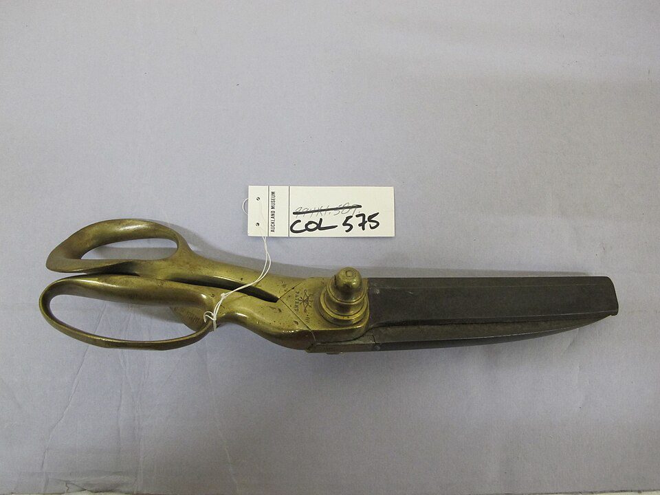 Antique industrial shears, brass handles with a golden patina, large steel blades, tagged with label reading "COL 575", laid on a light gray surface