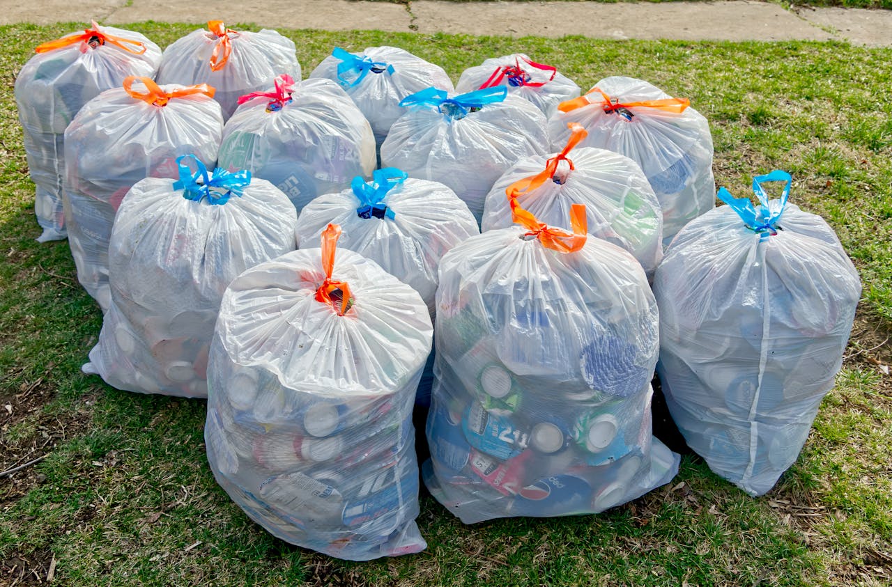Group of full white trash bags tied with blue, red, and orange ribbons, placed on grass near pavement