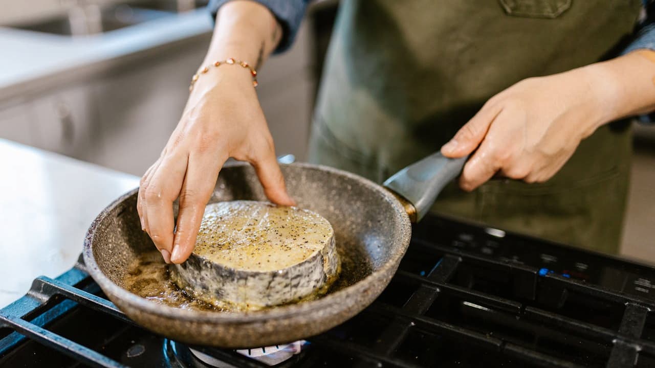 Hands placing a breaded food item into a frying pan, stovetop cooking, nonstick pan in use, wearing a green apron, kitchen background blurred.