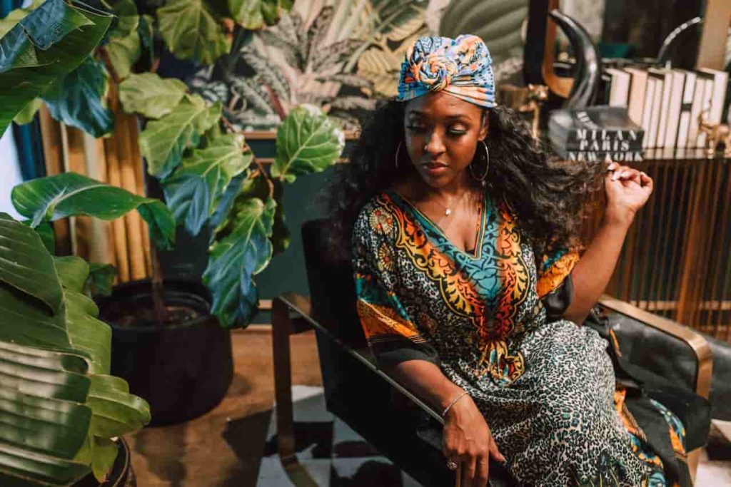 Woman wearing a colorful animal print dress and headwrap, sitting on a black chair, surrounded by large indoor plants, with patterned wallpaper and decor in the background