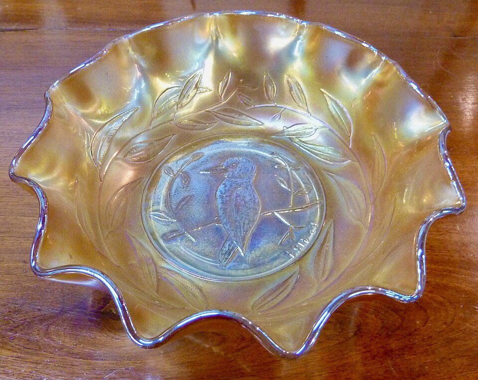 Iridescent carnival glass bowl, ruffled edge design, intricate embossed pattern at center, golden-yellow hue, displayed on wooden surface