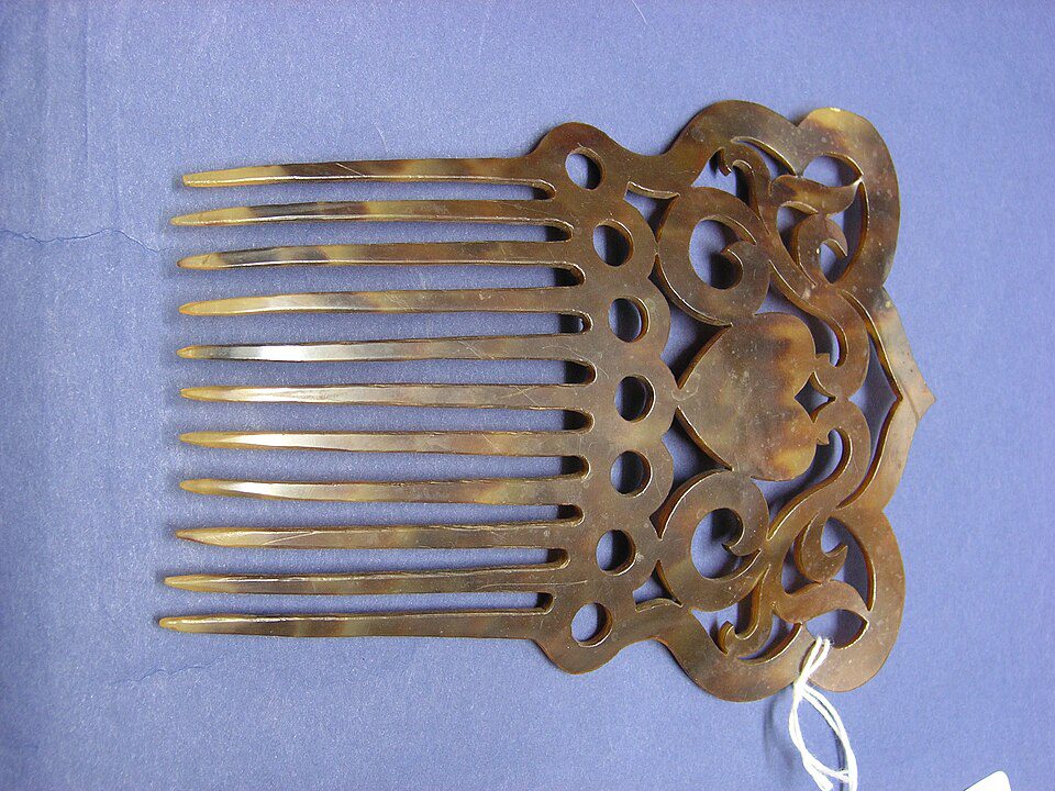 Antique decorative hair comb, brown tortoiseshell design, intricate carved floral pattern, wide teeth, placed on a blue surface, small white tag attached