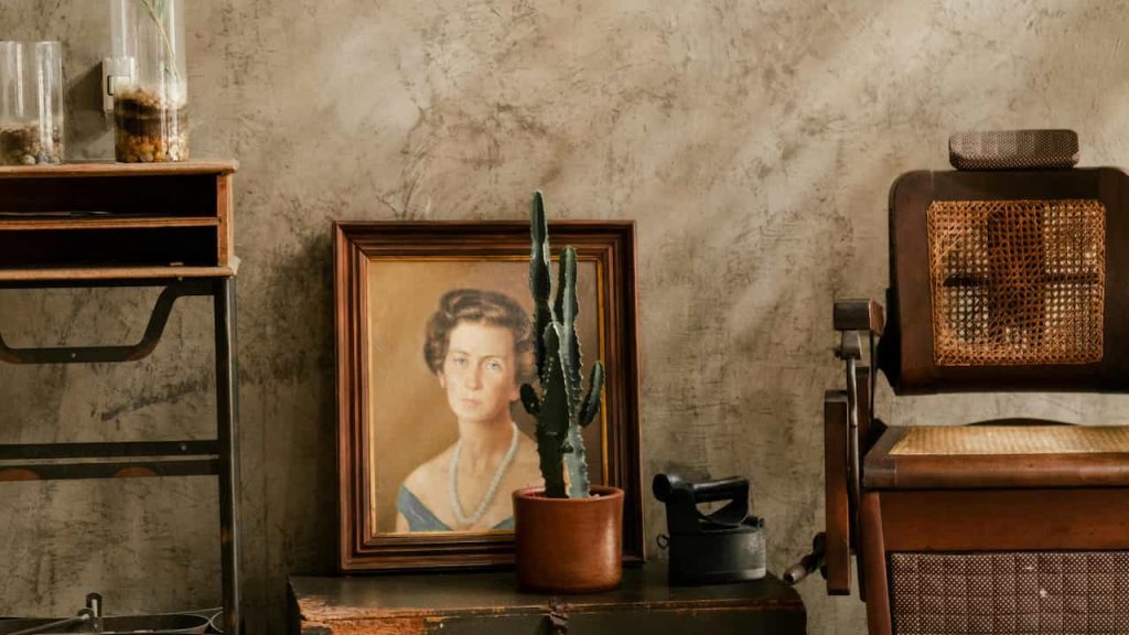 Framed portrait of a woman with a pearl necklace, leaning against a textured wall, cactus plant in a clay pot in front, antique wooden chair with cane backing, and old furniture surrounding the scene